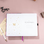 Open notebook featuring a gold foil 'M' logo and "This Journal Belongs To" page on a purple textured journal.