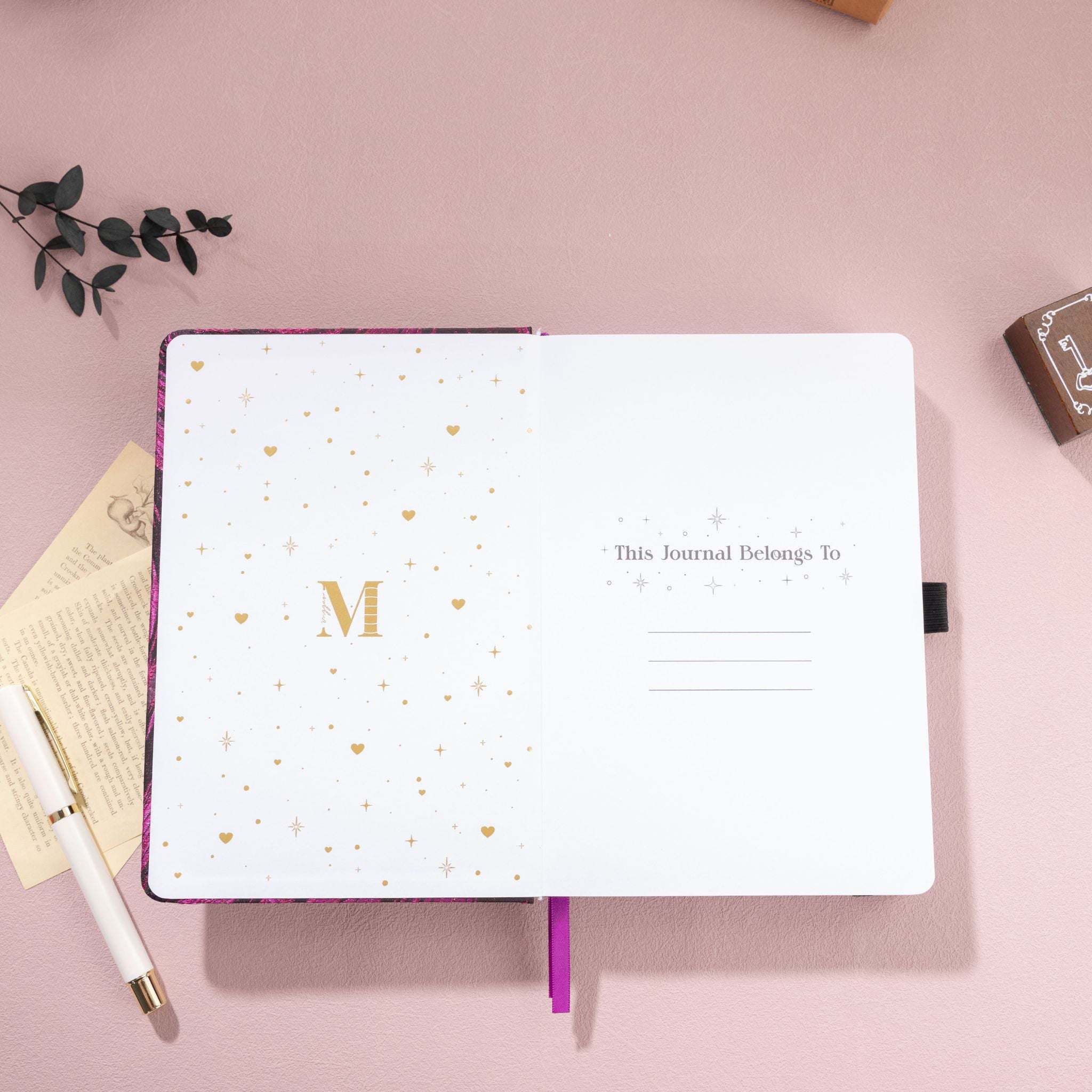 Open notebook featuring a gold foil 'M' logo and "This Journal Belongs To" page on a purple textured journal.