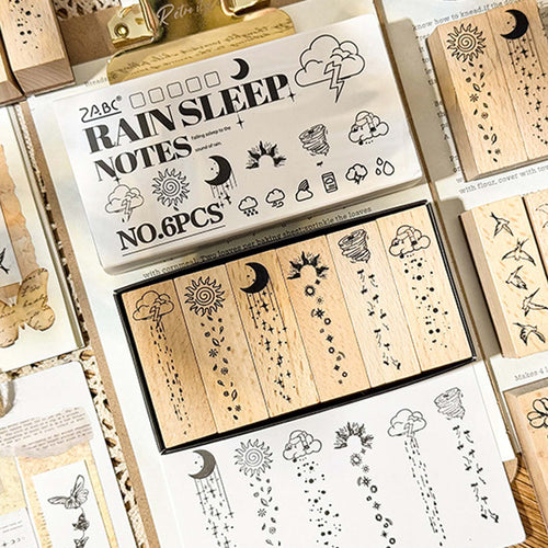 A flat lay of the Rain Sleep Notes wooden stamp set, showing the white packaging box with 6 long rectangular stamps inside, accompanied by stamped paper samples and loose wooden stamps on vintage handwritten paper.