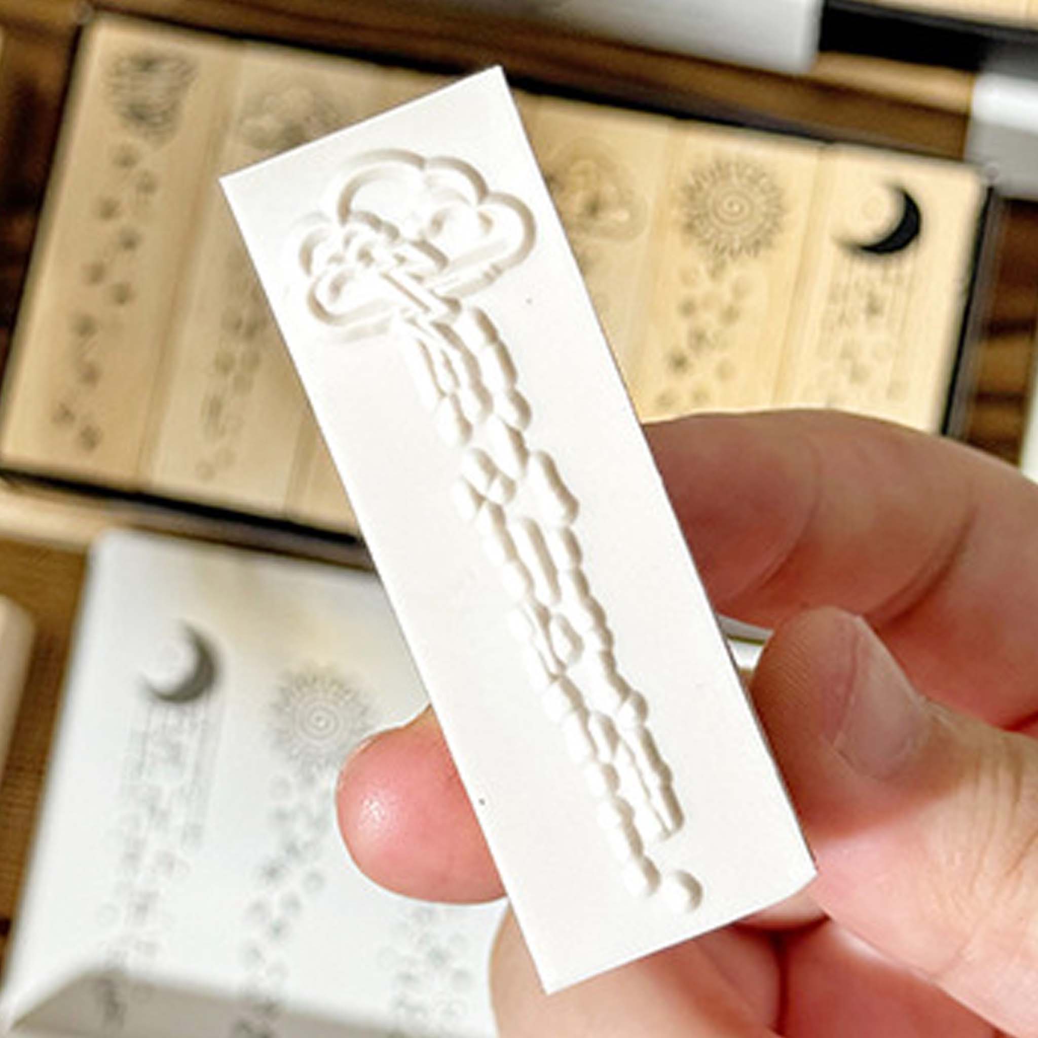 A hand holding a rectangular white rubber stamp surface from the Rain Sleep Notes set, showing an embossed cloud and raindrop design, with wooden stamps and packaging in the background.