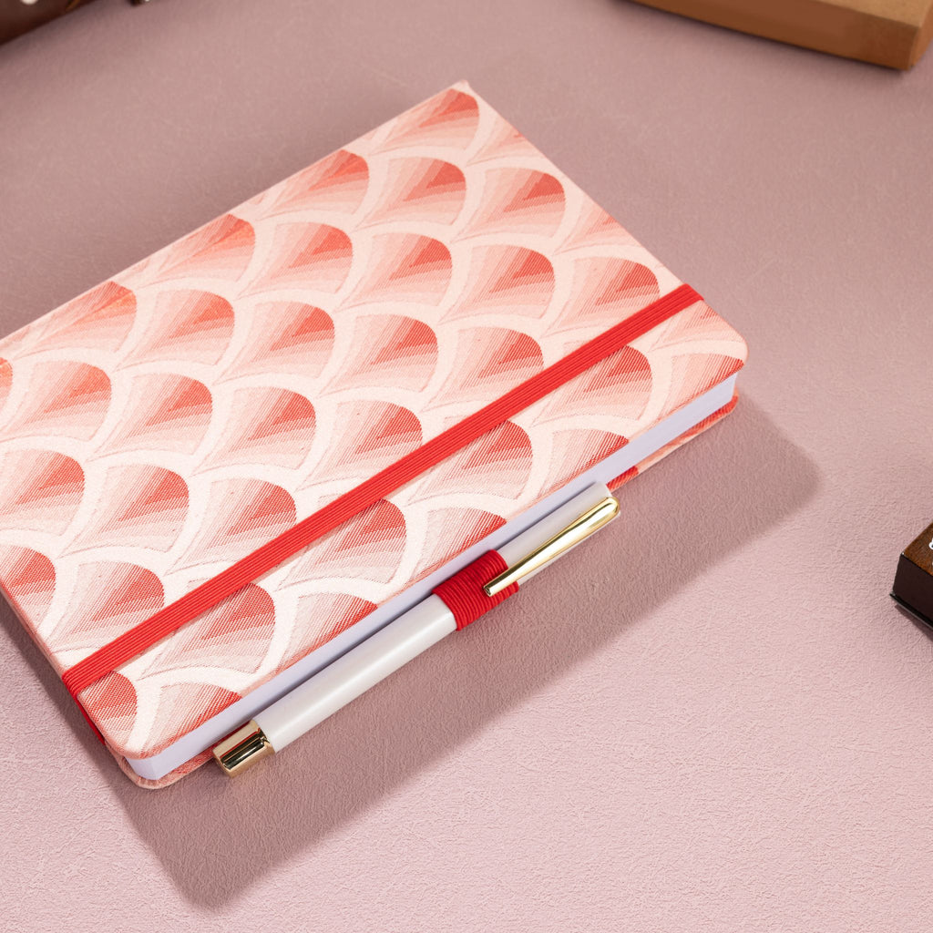 Red scalloped fabric notebook secured with a red elastic band, holding a white and gold pen