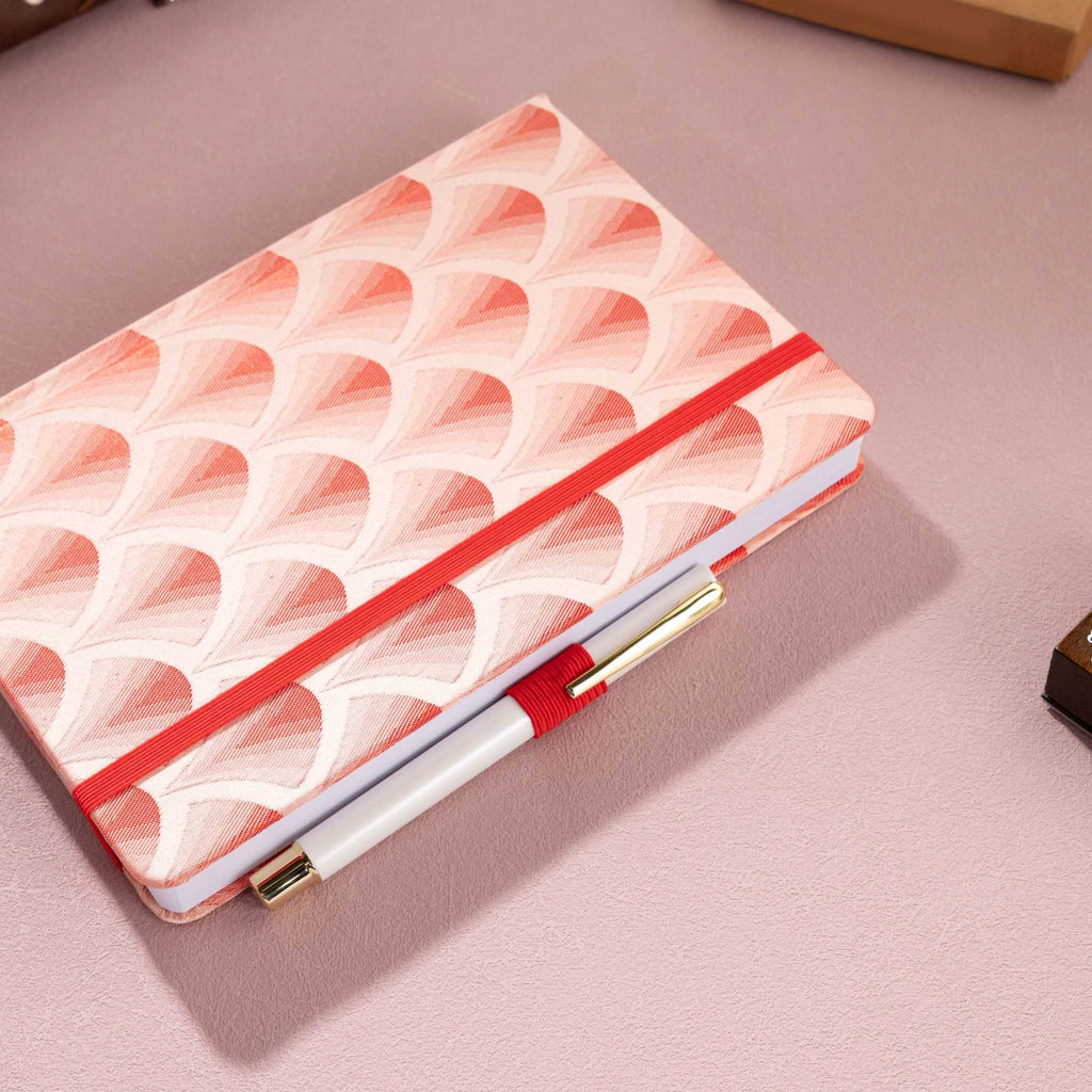 Red scalloped fabric notebook secured with a red elastic band, holding a white and gold pen