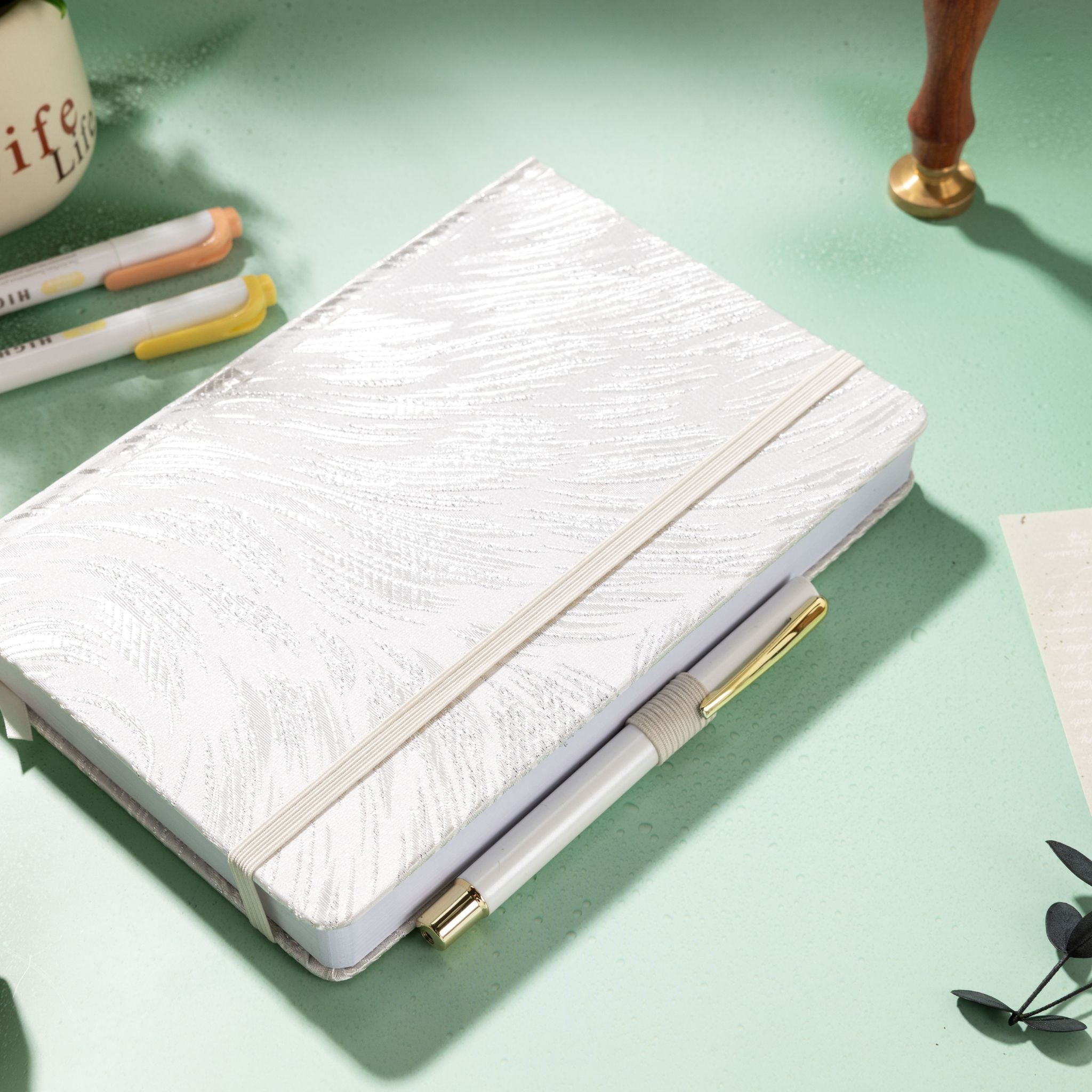 White and silver embroidered fabric notebook with elastic band, holding a white and gold pen