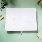 Inside title page featuring a gold foil 'M' logo and 'This Journal Belongs To' section on a silver embroidered journal