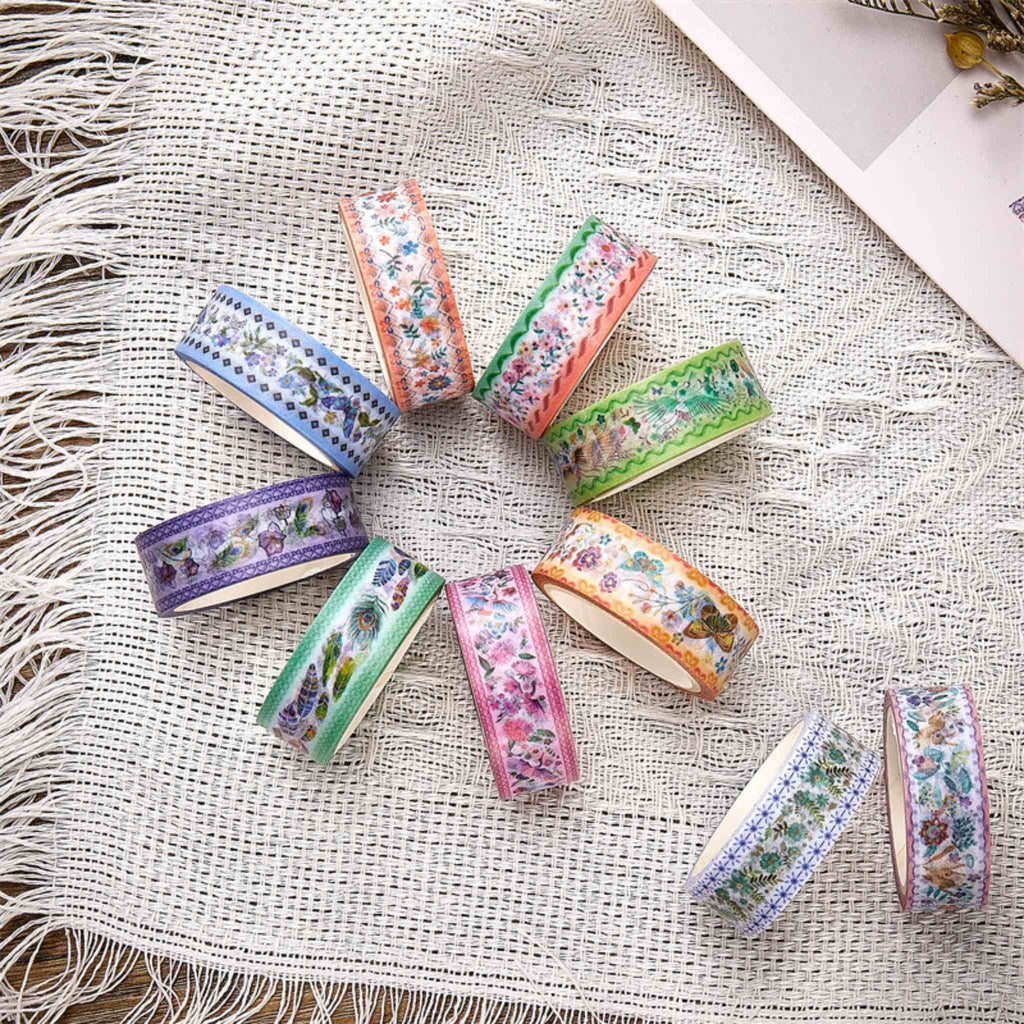A collection of vintage botanical and bird washi tapes arranged in a circle on a white fringed fabric, showing various pastel floral and feather motifs.