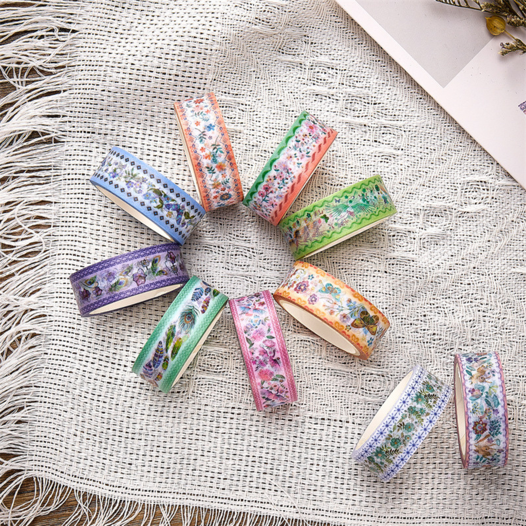 A collection of vintage botanical and bird washi tapes arranged in a circle on a white fringed fabric, showing various pastel floral and feather motifs.