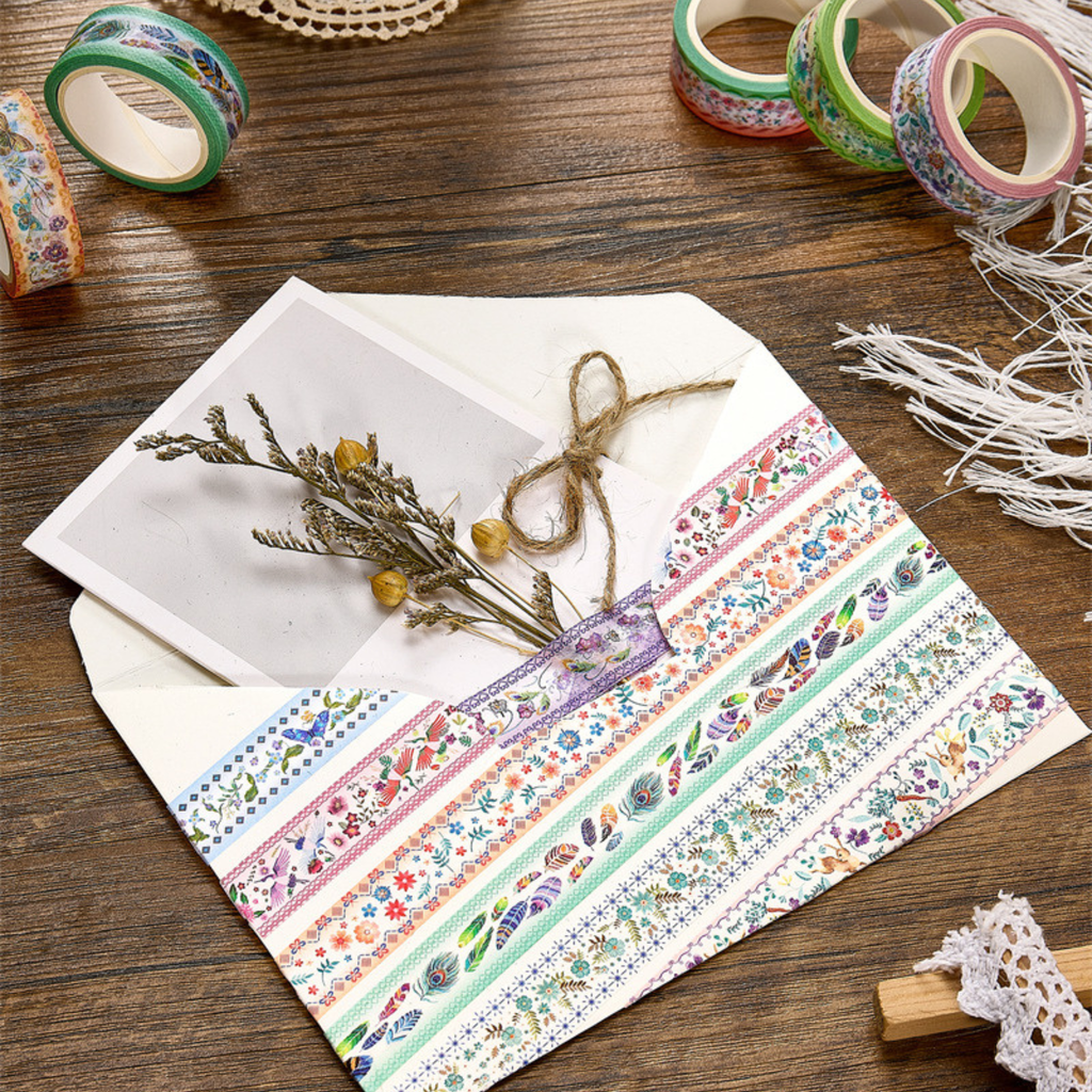 A white envelope decorated with vintage floral and bird washi tapes, tied with twine and paired with dried flowers, styled on a wooden surface.