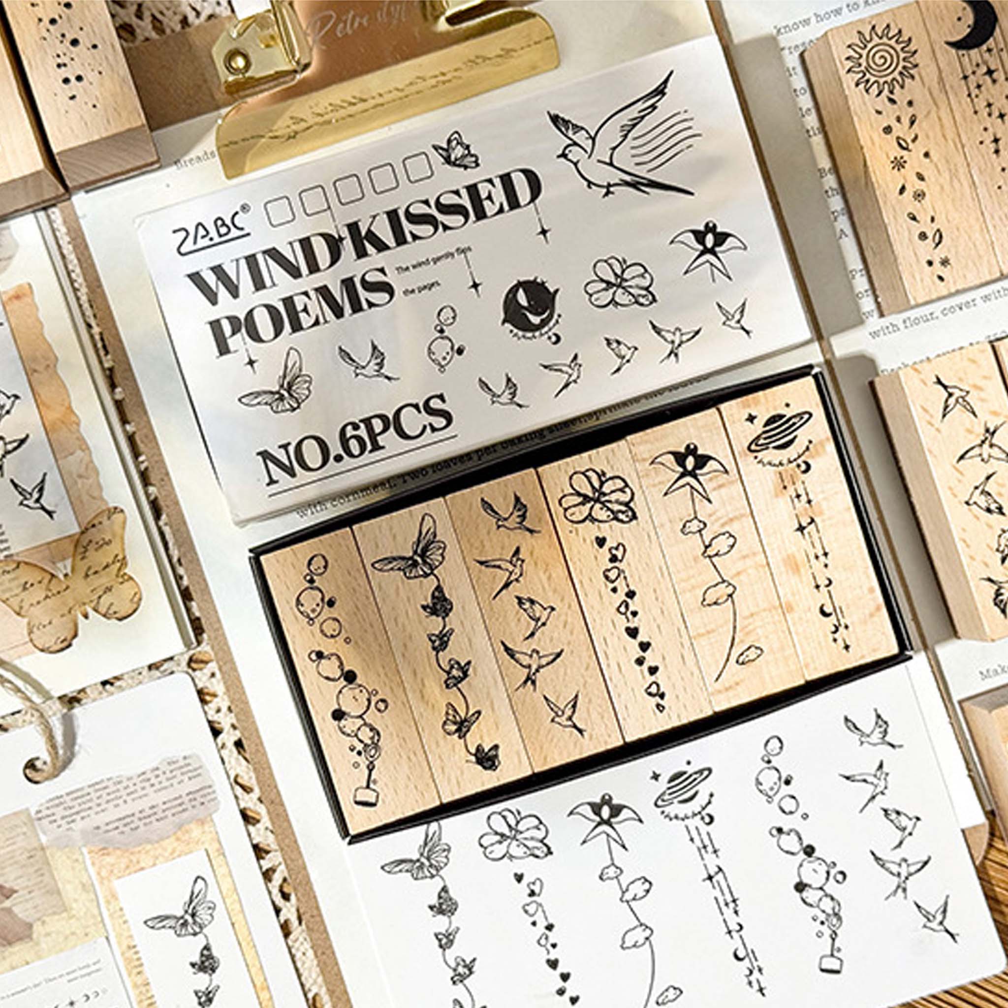 A flat lay of the Wind-Kissed Poems wooden stamp set, showing the white packaging box with 6 long rectangular stamps inside, along with stamped paper samples and loose stamps on vintage handwritten paper.