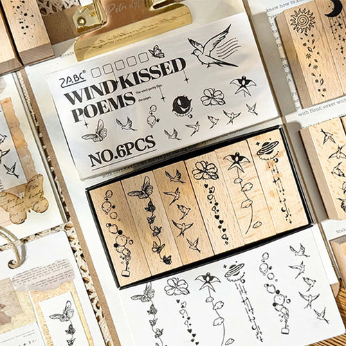 A flat lay of the Wind-Kissed Poems wooden stamp set, showing the white packaging box with 6 long rectangular stamps inside, along with stamped paper samples and loose stamps on vintage handwritten paper.
