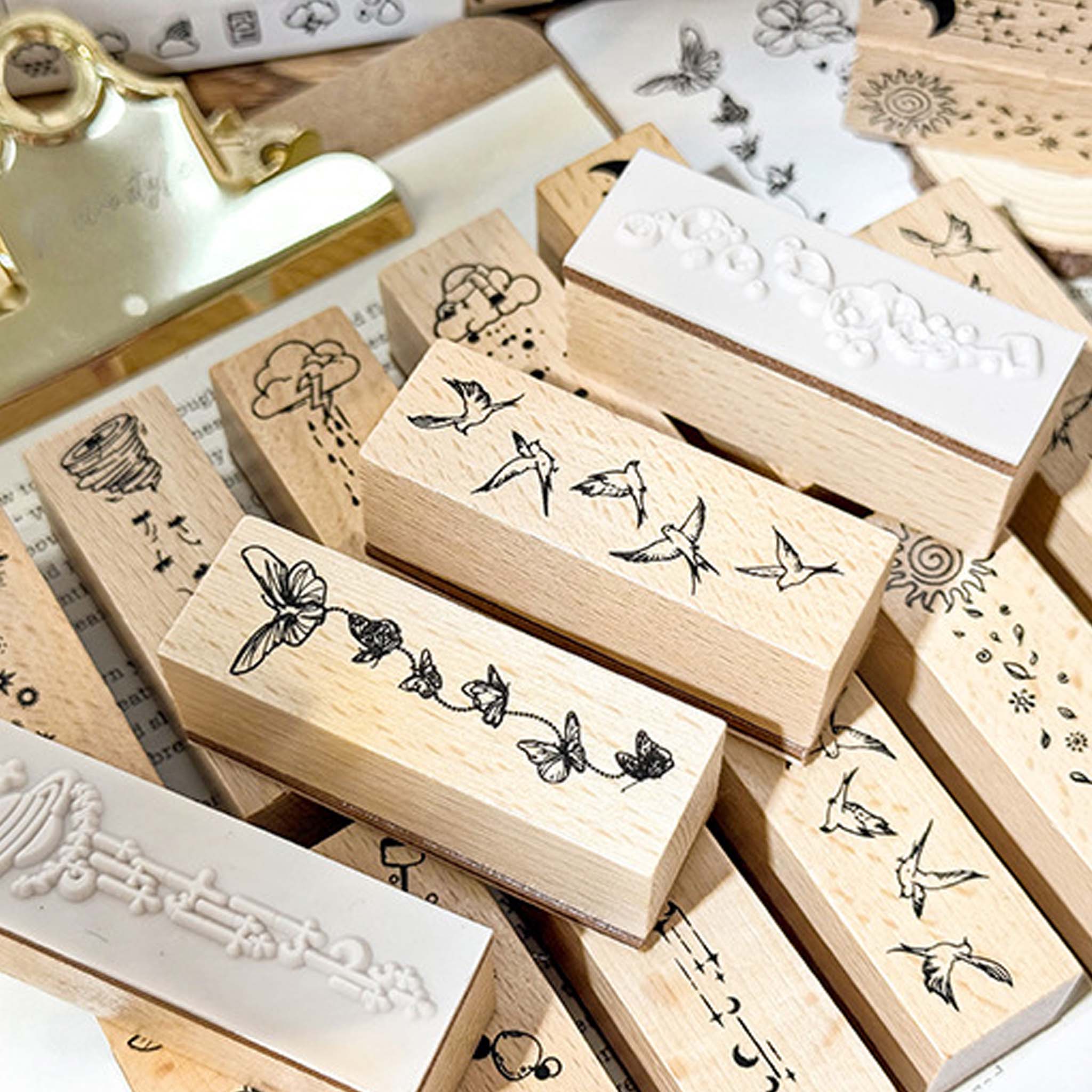 A close-up of wooden stamps from the Wind-Kissed Poems set, showing the natural wood grain, clear rubber stamping surfaces, and delicate line-art designs including butterflies, birds, clouds, and stars, arranged on vintage paper.