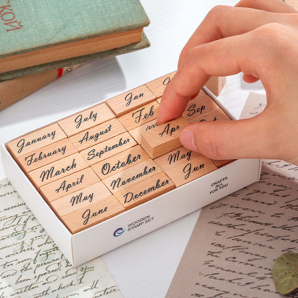 A hand picking up a wooden month stamp from a white storage box containing 24 wooden stamps with full and abbreviated month names in elegant cursive script, styled with vintage books and handwritten paper.