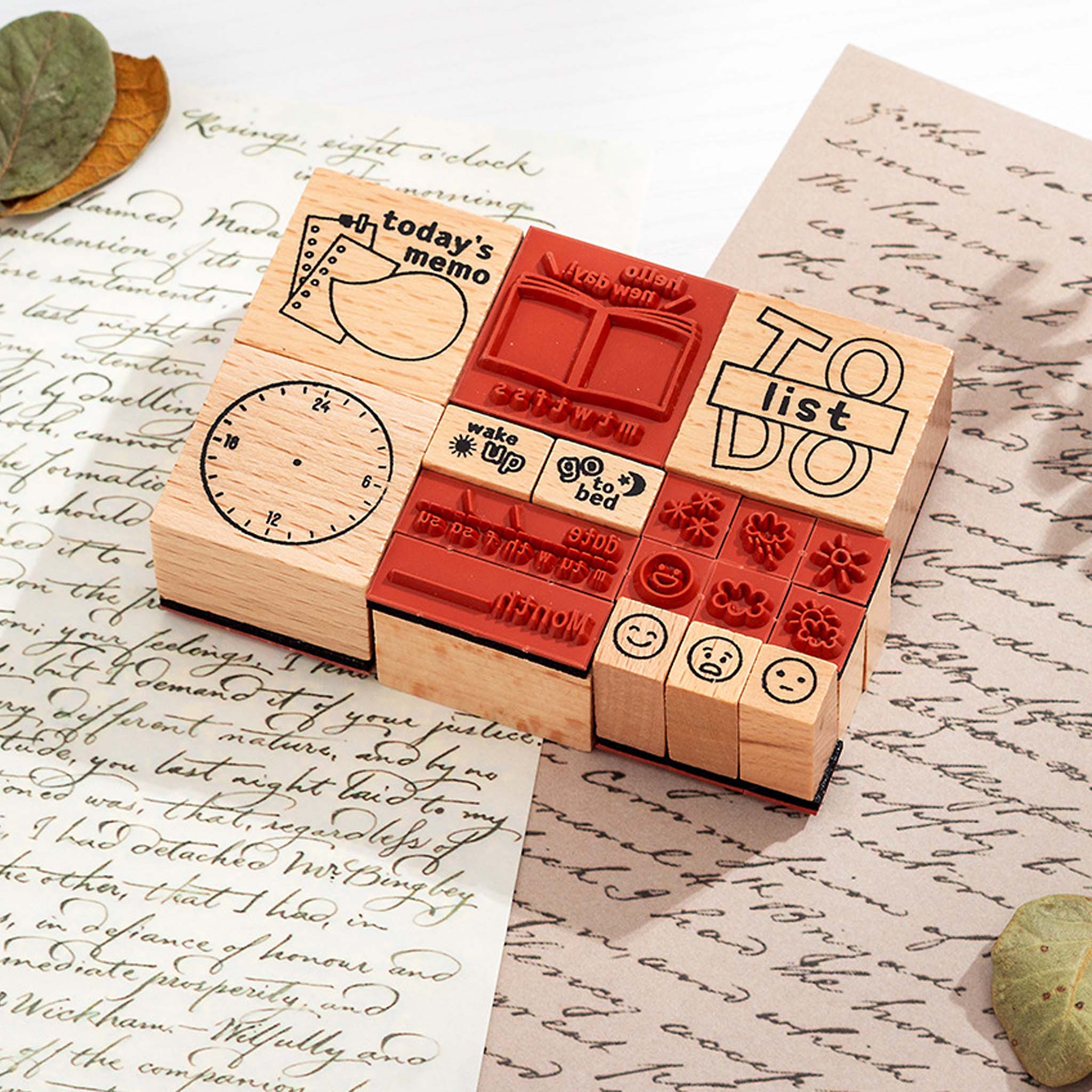 A close-up of wooden daily planner stamps with visible red rubber stamping surfaces, arranged on vintage handwritten paper with dried leaves, showcasing the stamp carving details and natural wood texture.