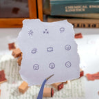 A torn piece of white paper stamped with 5 weather icons and 4 expression faces in purple ink, held by a pair of tweezers, with wooden stamps scattered in the background.