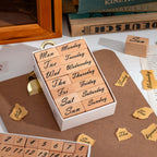 A white storage box containing 14 wooden weekday stamps, featuring both full and abbreviated forms of Monday through Sunday, arranged in a 2×7 grid on a white background.
