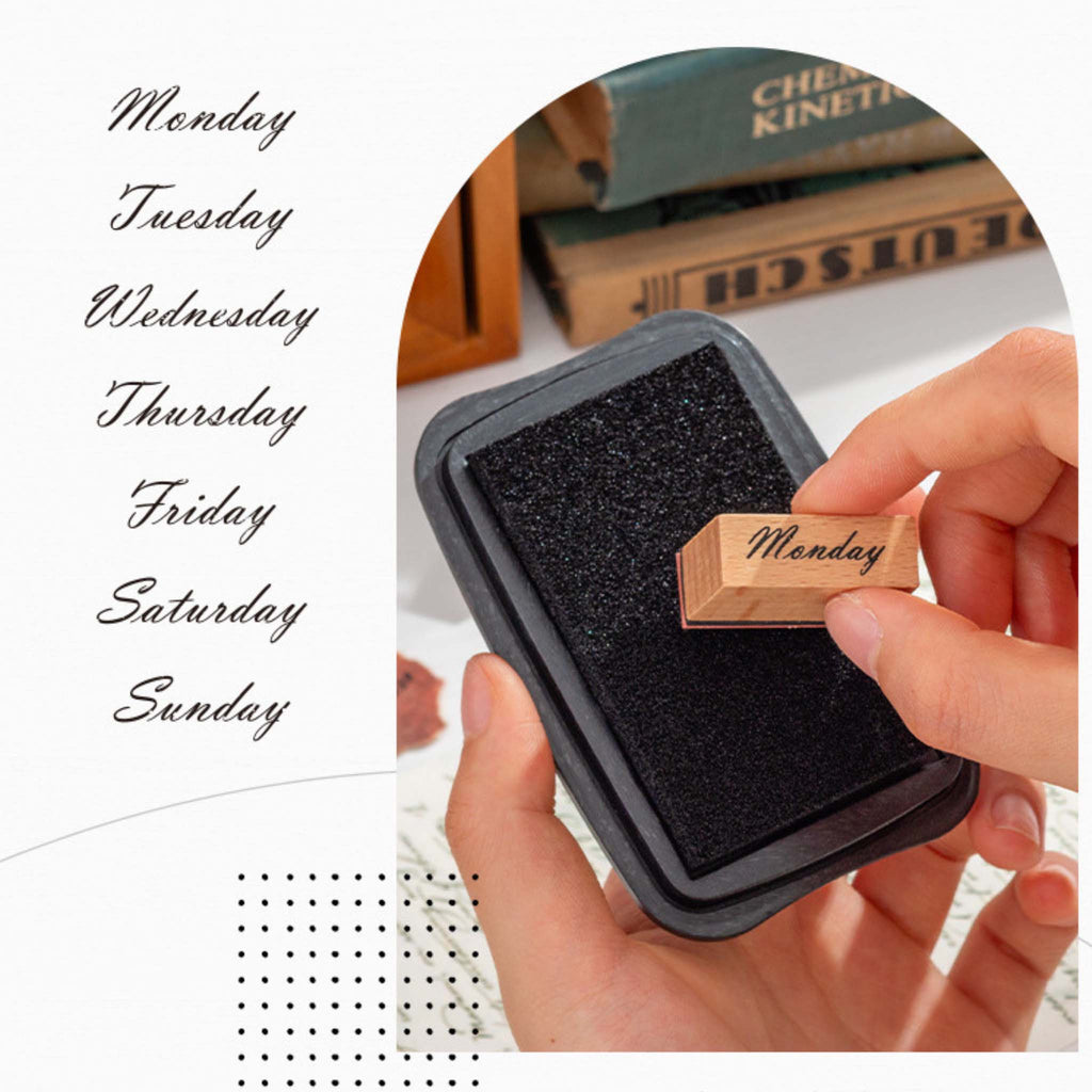 A hand pressing a wooden "Monday" stamp onto a black ink pad, with the stamp's natural wood grain and printed text clearly visible in a close-up shot.