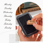 A hand pressing a wooden "Monday" stamp onto a black ink pad, with the stamp's natural wood grain and printed text clearly visible in a close-up shot.
